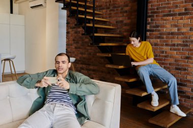 A young gay couple relaxes in their modern apartment, one on the couch scrolling on their phone, the other on the stairs using a laptop. clipart