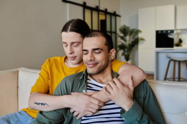 Genç eşcinsel bir çift modern bir dairede bir kanepede oturuyor, birbirlerine sarılıp eğleniyorlar..