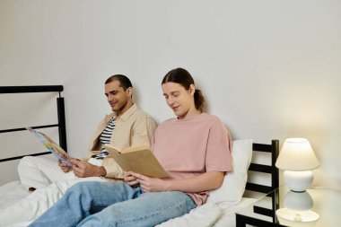 Gündelik giyinmiş iki genç adam modern bir dairede bir yatakta oturup kitap okuyor ve rahatlatıcı bir akşamın tadını çıkarıyor..