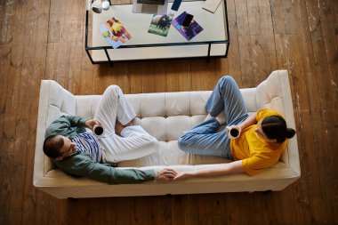 A young gay couple relaxes on a white sofa in a modern apartment, enjoying coffee together. clipart