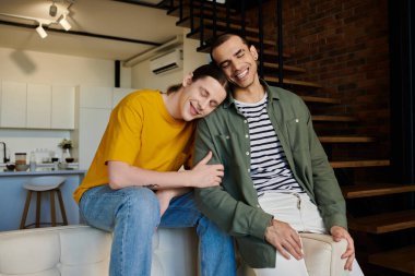 A young gay couple relaxes together on a white couch in their modern apartment, enjoying a quiet moment of intimacy. clipart