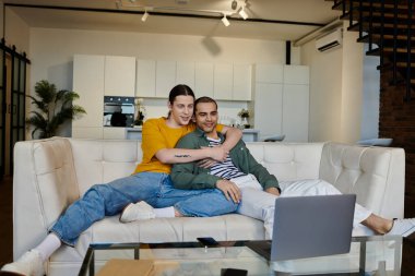 Genç eşcinsel bir çift, modern bir dairede dizüstü bilgisayarda film izlerken beyaz bir kanepeye sarılıyor..