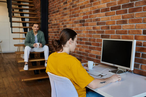 Молодая гей-пара наслаждается спокойным днем в своей современной квартире. Один партнёр работает за столом, а другой наблюдает с лестницы..