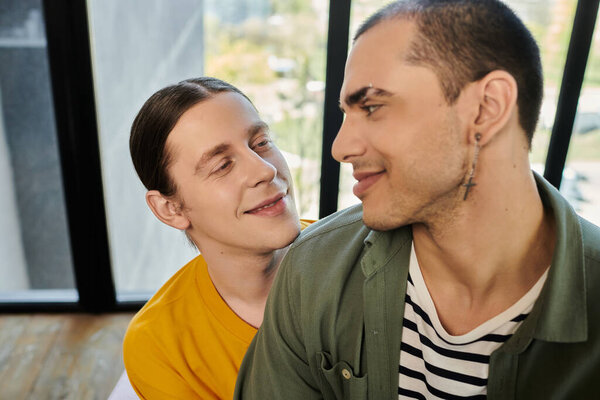 Two young gay men in casual attire share a loving look in a modern apartment.