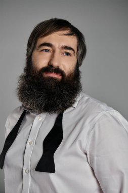Beyaz gömlekli, sakallı ve papyonlu bir adam gri arka planda rahatça poz veriyor..