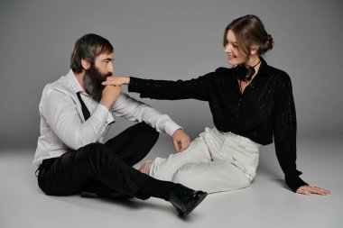 Sevgi dolu bir çift gri arka planda oturur, siyah bluzlu ve beyaz pantolonlu kadın, beyaz gömlekli ve siyah pantolonlu adam. Nazikçe eline dokunuyor..