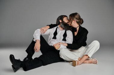 Resmi giyinmiş bir çift stüdyo zemininde otururken şefkatli bir öpücük paylaşıyor..