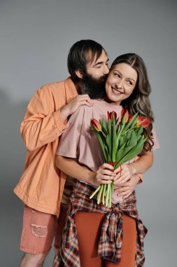 Sofistike giyinmiş bir kadın ve erkek stüdyo ortamında birlikte poz veriyorlar. Adam kadını yanağından öpüyor ve kadın elinde bir buket kırmızı lale tutuyor..