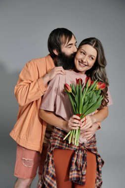 A man and woman in sophisticated attire pose together in a studio setting. The man kisses the woman on the cheek, and she holds a bouquet of red tulips. clipart