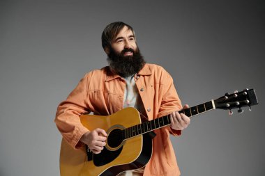 A man with a beard plays an acoustic guitar in a studio with a grey background.