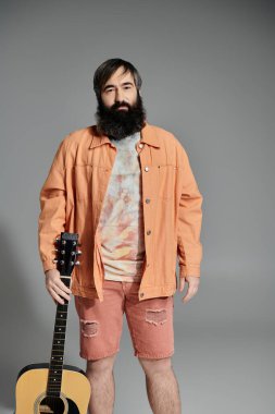 Elinde gitarla gri bir zemine karşı duran bir adam. Üzerinde mercan renkli bir ceket, şort ve kravat boyası bir gömlek var..