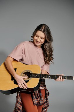 Pembe tişörtlü ve beline ekose gömlekli bir kadın gülümserken akustik gitar çalar..
