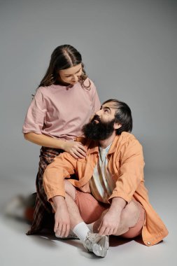 Bir çift stüdyo ortamında oturuyor, pembe gömlekli ve ekose pantolonlu bir kadın, turuncu ceketli ve şortlu bir adam, birbirine sevgiyle bakıyor..