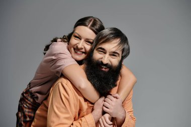 Bir kadın sakallı bir adama sarılıyor, ikisi de gri bir zemine gülümsüyor..