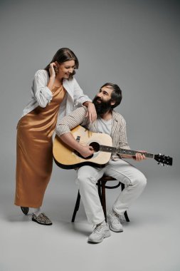 Bir erkek ve bir kadın stüdyo ortamında birlikte poz veriyorlar. O yanında dururken o akustik gitar çalıyor..