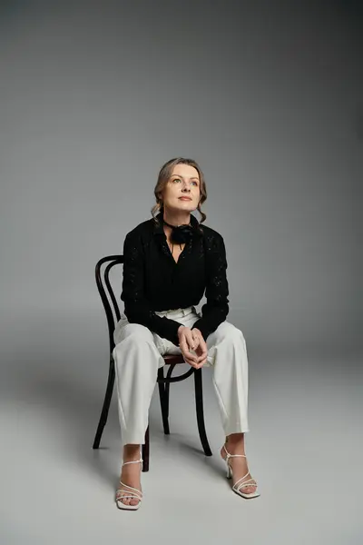 stock image A woman in a black blouse and white pants sits on a chair, gazing upwards.