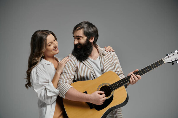 A man plays guitar while his partner rests her hand on his shoulder, both looking at each other with affection.