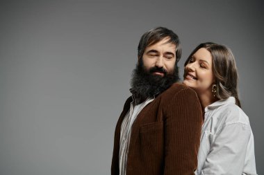 Birbirini seven bir çift, kahverengi kadife ceketli bir adam ve beyaz gömlekli bir kadın, düz gri bir zemine karşı poz verir..