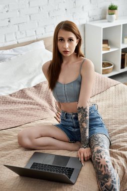 Dövmeli genç bir kadın evinde yatağında dizüstü bilgisayar kullanıyor..