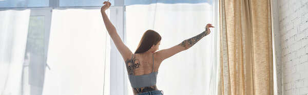 A young woman with tattoos stretches near a window in her modern apartment.