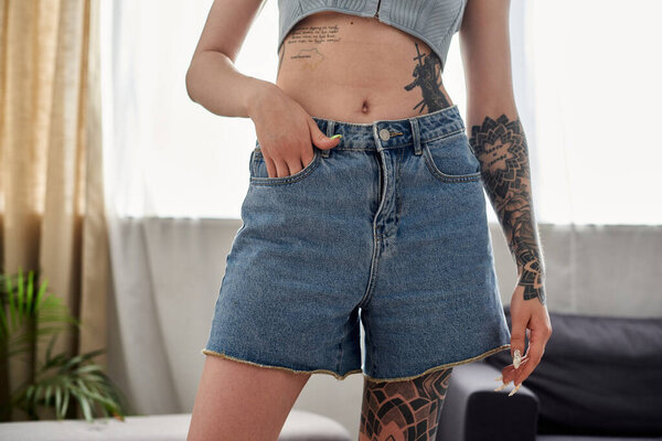 A young woman with tattoos on her arms and stomach stands in her modern apartment, wearing denim shorts.
