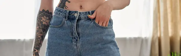 stock image A young woman with a tattoo on her arm stands in her modern apartment, wearing blue jeans and a relaxed expression.