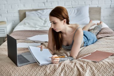 Dövmeleri olan genç bir kadın evde laptopuyla çalışırken yatakta yatıyor..