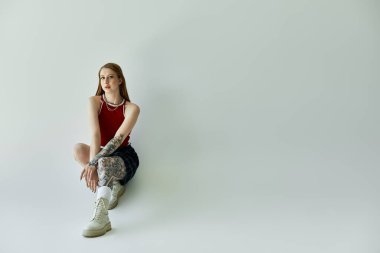 A young woman with tattoos sits with her legs crossed against a plain white background. She wears a red tank top, a black and white plaid skirt, and white boots. clipart