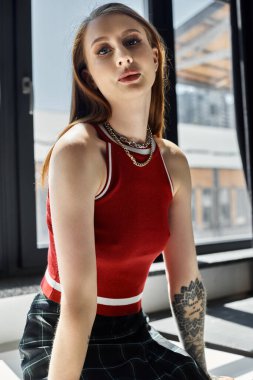 A young woman in a red tank top and plaid skirt sits by a window, her tattoo visible. clipart