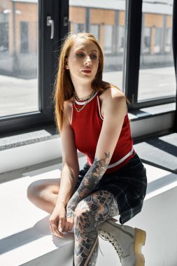 Dövmeleri olan genç bir kadın beyaz bir çıkıntıda oturuyor kırmızı bir atlet ve ekose etek giyiyor..