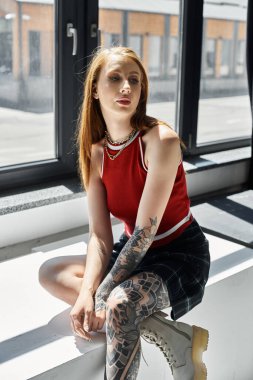 A young woman with tattoos sits on a white ledge wearing a red tank top and a plaid skirt. clipart