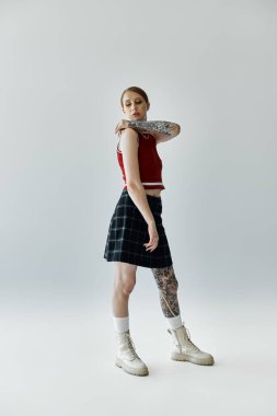 Dövmeli genç bir kadın gri bir arkaplanın önünde duruyor, kırmızı bir bluz giyiyor, siyah beyaz bir etek ve beyaz çizmeler giyiyor..