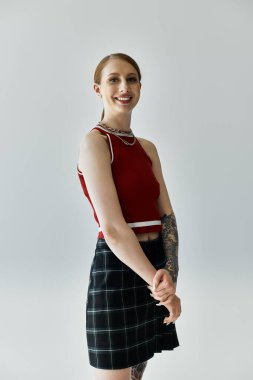 Dövmeleri olan genç bir kadın, gri bir arkaplanda siyah beyaz bir etek ve kırmızı bir atlet ile poz veriyor..