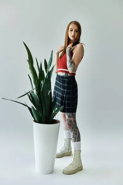 stock image A tattooed young woman wearing a plaid skirt and a red tank top stands next to a large green plant in a white pot.