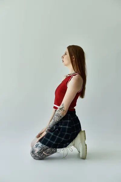 stock image A young woman with tattoos on her arms and legs poses in a plaid skirt and red tank top against a grey backdrop.