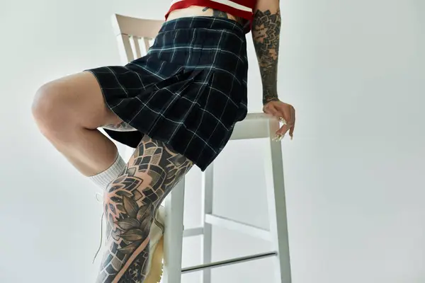 stock image A tattooed young woman poses in a plaid skirt and tank top against a grey background, showcasing a blend of youthfulness and fashion.