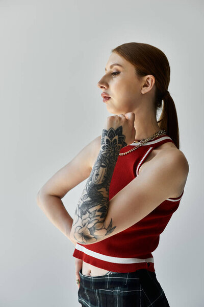 A young woman with a full sleeve tattoo stands against a grey background, wearing a red tank top and plaid skirt.