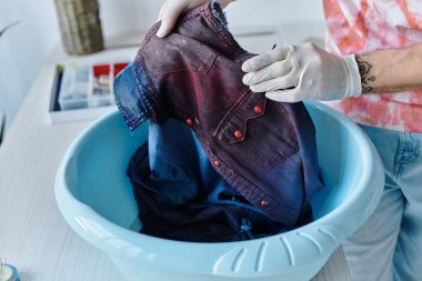 Kravat boyalı gömlekli genç bir adam mavi plastik bir leğende kot gömleğini yıkıyor, sürdürülebilir DIY yöntemleriyle atılmış giysileri canlandırıyor..