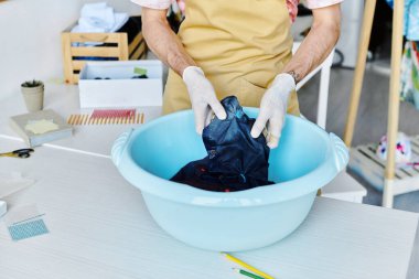 Önlüklü ve eldivenli genç bir adam, sürdürülebilir moda yükseltme işinin bir parçası olarak leğene bir giysi boyuyor..