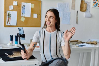 A young man with long hair, wearing a striped shirt and a rainbow tattoo, smiles and waves while recording a video in his DIY clothing restoration atelier. clipart