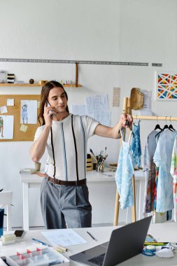 DIY giysi restorasyon atölyesinde genç, eşcinsel bir adam telefonda, sürdürülebilir giyim markası için yeniden tasarlanmış bir elbise sergiliyor..