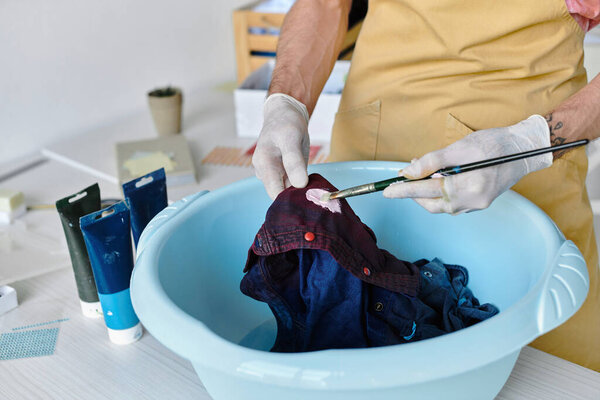 A young man paints a discarded shirt with a brush in a basin of water, revitalizing it in his DIY clothing restoration atelier.