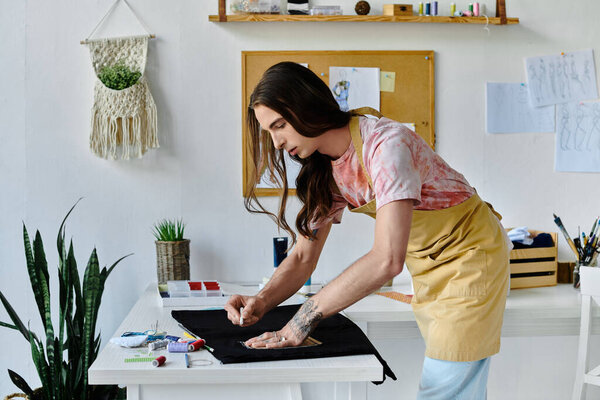 A young man, with long hair and a tie-dye shirt, is working on a piece of clothing in his home studio, focused on giving new life to discarded items.