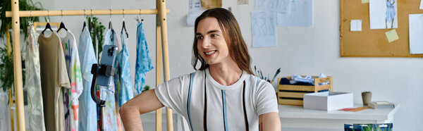 A young man smiles as he works in his clothing restoration atelier, surrounded by racks of vibrant fabrics and a phone camera for capturing his creations.