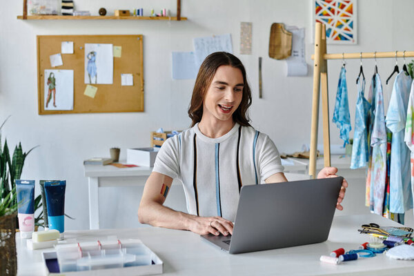 A young gay man works on his laptop in his clothing restoration atelier, surrounded by fabrics and tools. He focuses on giving old clothes new life.