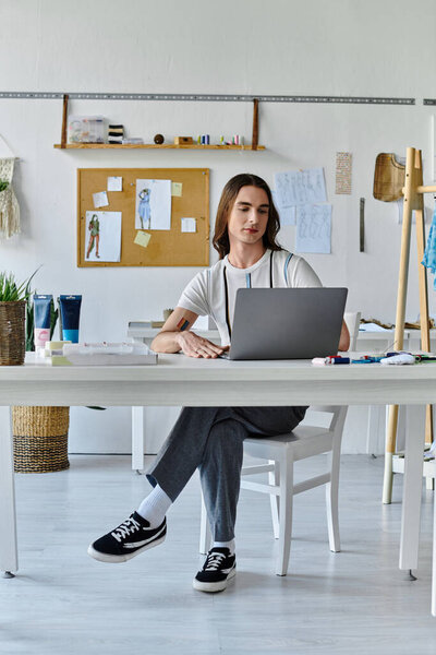 A young man sits at his desk in his clothing restoration atelier, working on his laptop. He is surrounded by tools and supplies for his creative work.