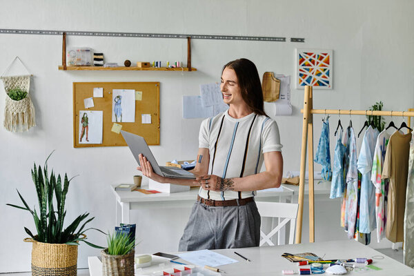 Молодой человек улыбается, когда использует свой ноутбук в мастерской по реставрации одежды. Он фокусируется на том, чтобы дать новую жизнь выброшенной одежде и продвигать устойчивые методы моды.