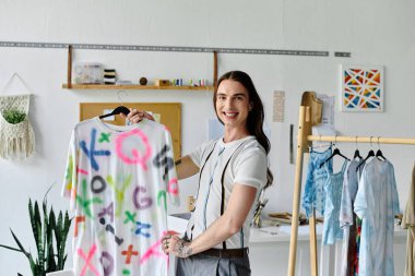 Genç bir adam, yenilenmiş bir tişörtü giysi yenileme atölyesinde sergilerken gururla gülümsüyor ve sürdürülebilir moda tutkusunu sergiliyor..