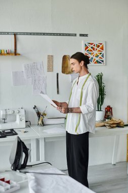 Beyaz gömlekli genç bir adam atölyesinde dikim malzemeleri ve dikiş makinesiyle çevrili bir tasarımı inceliyor..