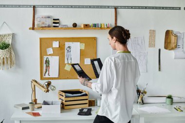Beyaz gömlekli genç bir adam, elinde kumaş örnekleri olan, DIY giysi restorasyon atölyesinde duruyor. Sürdürülebilirliğe olan bağlılığını gösteriyor ve eski kıyafetlere yeni bir hayat veriyor..